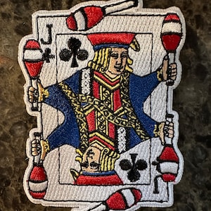 Puede incluir: Un parche bordado blanco del Jack of Clubs de la baraja de cartas. El Jack está sosteniendo palos de malabares en cada mano. Los palos son rojos, blancos y azules.