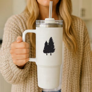 May include: A tall white insulated tumbler with a handle, a clear lid, and a clear straw. The front of the tumbler features a black graphic silhouette of two evergreen trees. The cup has a tapered base and a smooth, matte-textured finish.