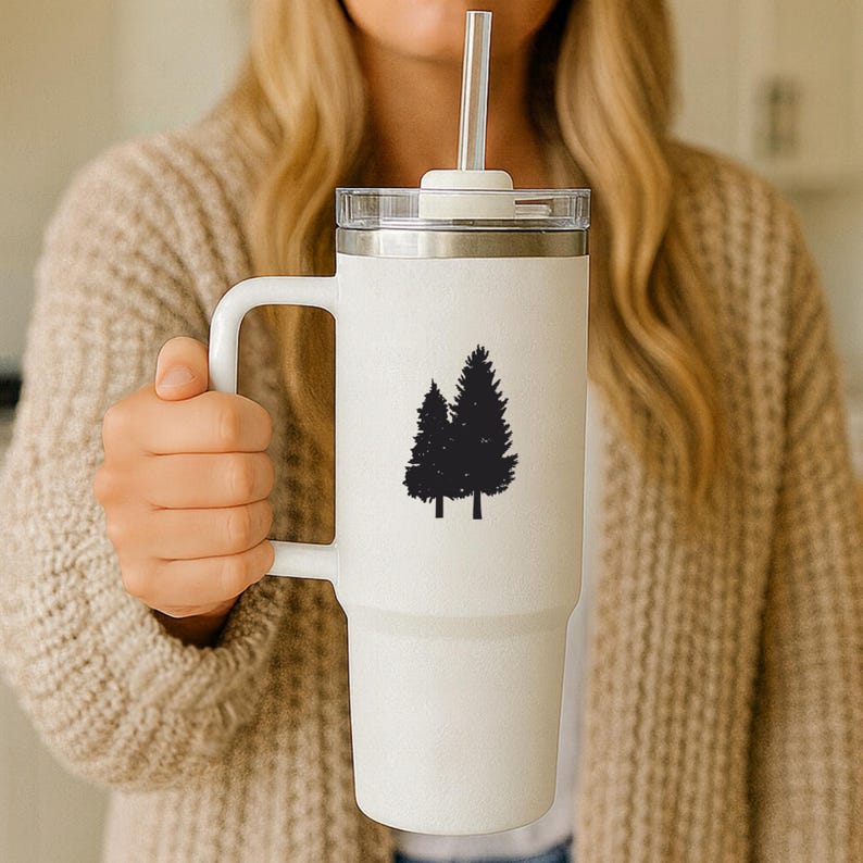 May include: A tall white insulated tumbler with a handle, a clear lid, and a clear straw. The front of the tumbler features a black graphic silhouette of two evergreen trees. The cup has a tapered base and a smooth, matte-textured finish.