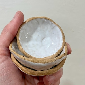 May include: Two small, round, handmade ceramic bowls stacked together. Each bowl features a textured, unglazed tan clay exterior rim and a glossy, uneven white glazed interior surface with visible indentations and a rustic, organic shape.