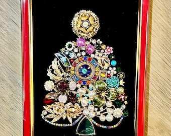 May include: A framed artwork featuring a Christmas tree design made from various colorful vintage jewelry pieces. The tree is set against a black background within a red and gold frame. The jewelry includes brooches, beads, and rhinestones.