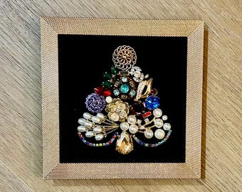 May include: A framed artwork featuring a Christmas tree design made from various vintage brooches and jewelry. The tree is composed of colorful gemstones, pearls, and gold-toned metal pieces, set against a black velvet background within a gold frame.