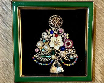 May include: A framed, jeweled Christmas tree design. The tree is composed of various colorful gemstones, pearls, and floral elements, set against a black background. The frame is a vibrant green with gold trim.