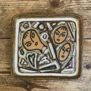 May include: A square ceramic wall plaque featuring a textured surface with three stylized faces outlined in dark brown. The design incorporates abstract geometric shapes and lines in shades of cream, light green, and dark brown, with a thick brown border.
