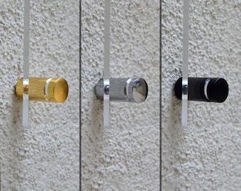 Pode incluir: Três puxadores de porta cilíndricos em ouro, prata e preto, cada um com um detalhe metálico a condizer. Os puxadores são montados numa parede texturizada de cor clara, com uma barra vertical transparente a estender-se de cada um.