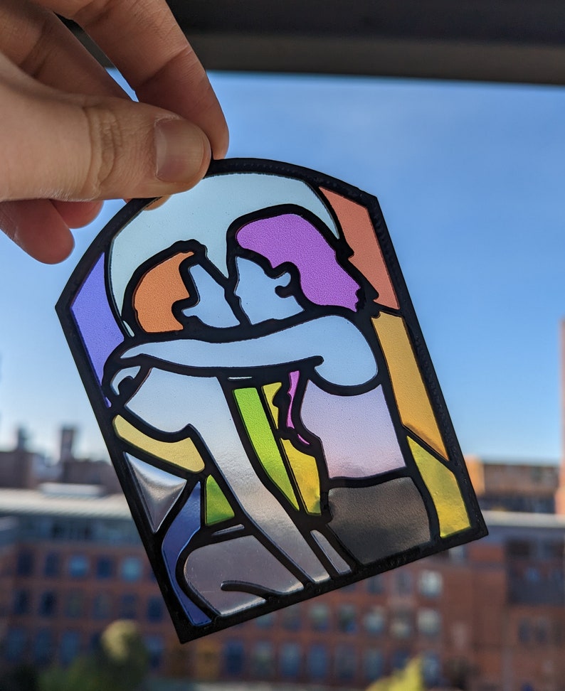 May include: A stained glass window-style design featuring two figures embracing. The figures are outlined in black and filled with various colors, including purple, orange, yellow, and green.
