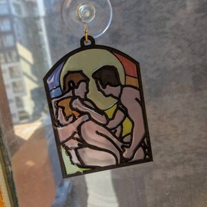 May include: A stained glass window hanging ornament featuring a couple embracing. The couple is depicted in a stylized, abstract style with a yellow, orange, and blue background.