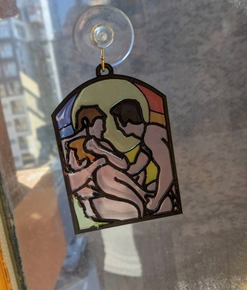 May include: A stained glass window hanging ornament featuring a couple embracing. The couple is depicted in a stylized, abstract style with a yellow, orange, and blue background.