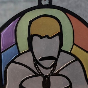Puede incluir: Un colgante de estilo vitral con una persona con un halo de arcoíris. La persona lleva un collar de cadena y tiene un fondo gris oscuro.
