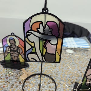 May include: A stained glass style image of two people embracing, with a rainbow color scheme. The image is mounted on a black metal stand with a circular base.