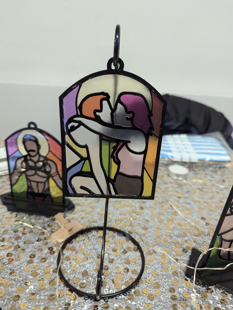 May include: A stained glass style image of two people embracing, with a rainbow color scheme. The image is mounted on a black metal stand with a circular base.