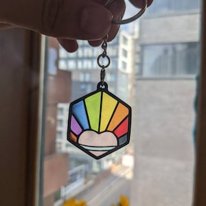 May include: A rainbow-colored hexagonal keychain with a white heart in the center. The keychain is attached to a silver ring and chain.