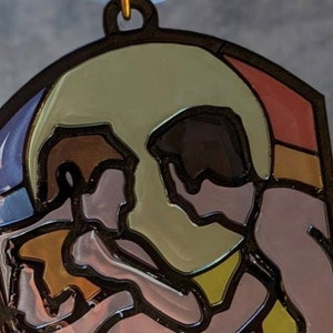 May include: A stained glass style ornament featuring two figures embracing, with a large, round, yellow-toned shape behind them. The figures are outlined in black and have a pink and brown color scheme. The background is a mix of blue, red, and orange.