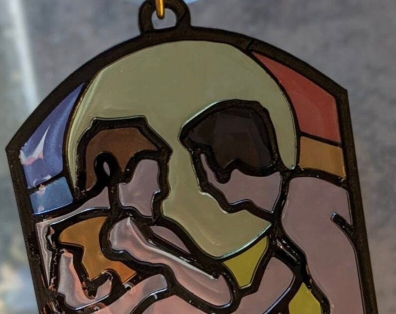 May include: A stained glass style ornament featuring two figures embracing, with a large, round, yellow-toned shape behind them. The figures are outlined in black and have a pink and brown color scheme. The background is a mix of blue, red, and orange.