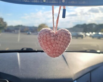 May include: A pink crocheted heart hanging from the rearview mirror of a car. The heart is fuzzy and has a textured surface.