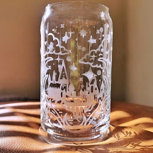 Może przedstawiać: Przezroczysty szklany kubek z białym grawerowanym wzorem zamku, chmur i gwiazd. Kubek stoi na brązowej powierzchni.