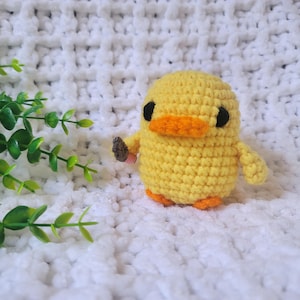 May include: A yellow crocheted duck with black eyes and an orange beak. The duck is sitting on a white fluffy surface.