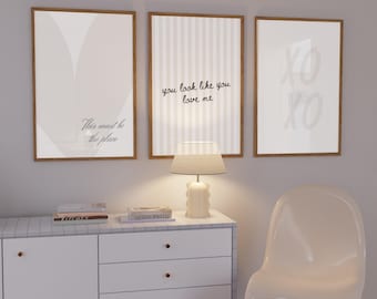 May include: Three framed art prints with wooden frames hang on a grey wall. The prints feature minimalist designs and text: "This must be the place," "you look like you love me," and "XO XO." A white lamp sits on a white cabinet.