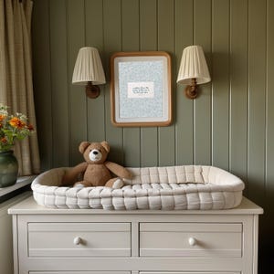 Puede incluir: Una escena de guarder&iacute;a con un cambiador blanco sobre una c&oacute;moda blanca y un oso de peluche marr&oacute;n. Dos apliques de pared con pantallas beige y una impresi&oacute;n enmarcada con texto.