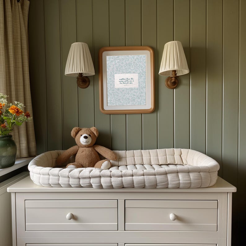 Puede incluir: Una escena de guarder&iacute;a con un cambiador blanco sobre una c&oacute;moda blanca y un oso de peluche marr&oacute;n. Dos apliques de pared con pantallas beige y una impresi&oacute;n enmarcada con texto.