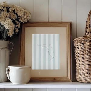 May include: A framed print featuring a black outline of the state of Florida on a light blue and white striped background. The print is in a wooden frame and is displayed on a white shelf with a white vase and a wicker basket.