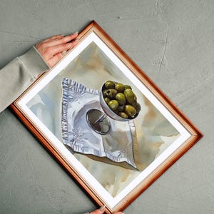 May include: A framed watercolor painting depicts a still life of green olives in a silver goblet on a crumpled white napkin. The painting is set against a neutral background, and the frame is a warm brown color.