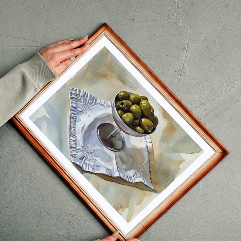 May include: A framed watercolor painting depicts a still life of green olives in a silver goblet on a crumpled white napkin. The painting is set against a neutral background, and the frame is a warm brown color.