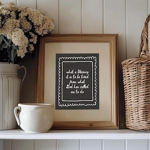 May include: A framed print with the text "what a blessing it is to be tired from what God has called me to do." The print is in a wooden frame and is displayed on a white shelf with a white vase of flowers and a woven basket.
