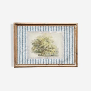 Op de afbeelding: Een ingelijste print met een aquarel schilderij van een grote boom met groen en geel blad. Het kunstwerk is geplaatst tegen een blauw-wit gestreepte stof, omlijst in een houten frame.