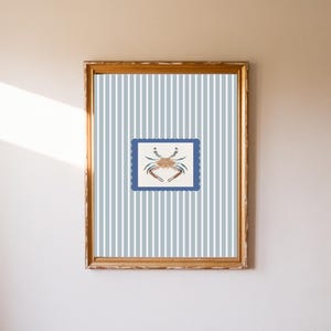 May include: A framed print featuring a crab illustration. The crab is depicted in shades of brown, beige, and blue, set against a blue and white striped background. The frame is gold-toned, adding a touch of elegance to the coastal-themed artwork.