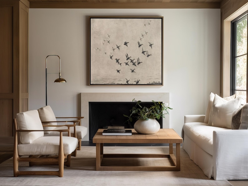 May include: A living room scene featuring a square framed artwork depicting birds in flight above a landscape. The room includes a fireplace, two armchairs, a sofa, and a wooden coffee table with a vase and books. A brass floor lamp provides additional lighting.