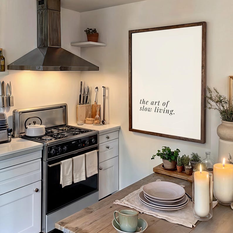 May include: A kitchen interior with a stainless steel extractor fan, cooker, and a wooden dining table. A framed print on the wall displays the text "the art of slow living." Candles and plants create a warm ambiance.