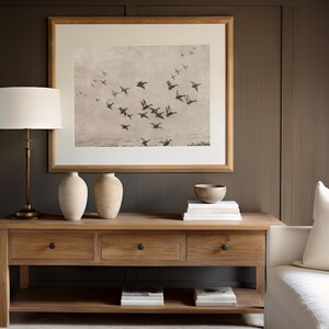 May include: A wooden console table with three drawers and a lower shelf, holding books. A framed print of flying birds hangs above. Two cream-colored vases, a small bowl, and a lamp with a white shade are on the table. The wall is a dark brown color.