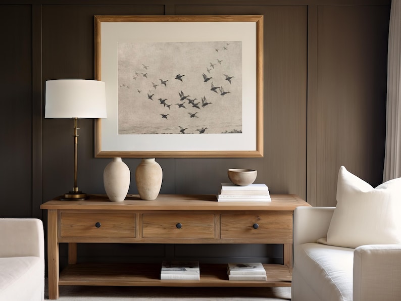 May include: A wooden console table with three drawers and a lower shelf, holding books. A framed print of flying birds hangs above. Two cream-colored vases, a small bowl, and a lamp with a white shade are on the table. The wall is a dark brown color.