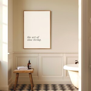 May include: A bathroom interior with a framed print reading "the art of slow living." A small wooden stool supports a rolled white towel and a brown bottle. The floor is patterned with black and white tiles.