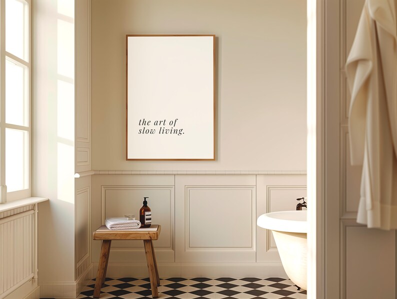 May include: A bathroom interior with a framed print reading "the art of slow living." A small wooden stool supports a rolled white towel and a brown bottle. The floor is patterned with black and white tiles.