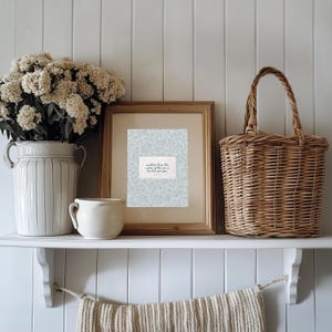 Puede incluir: Una estanter&iacute;a blanca exhibe una impresi&oacute;n enmarcada con el texto "mightier than the waves of the sea is his love for you." Un jarr&oacute;n blanco con flores, una taza peque&ntilde;a y una cesta de mimbre completan el arreglo.