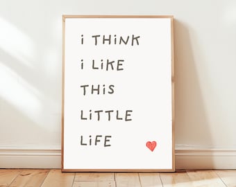 May include: A framed print featuring the text "i THINK i LIKE THIS LITTLE LIFE" in a handwritten font, with a small red heart. The frame is light brown. The print is displayed against a white wall.