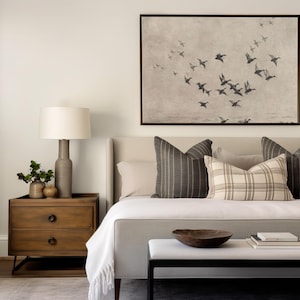 May include: A bedroom scene featuring a bed with decorative pillows, a wooden nightstand with a lamp, and a framed artwork depicting birds in flight. The room has a neutral color palette.