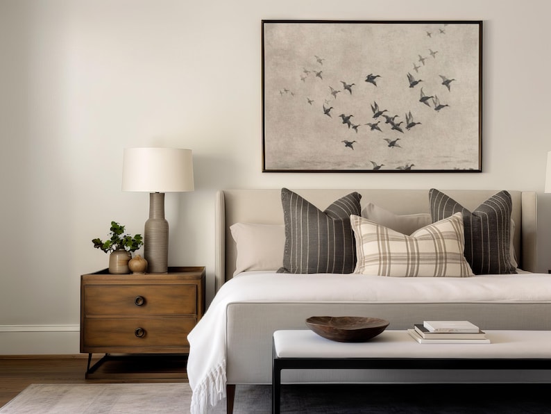 May include: A bedroom scene featuring a bed with decorative pillows, a wooden nightstand with a lamp, and a framed artwork depicting birds in flight. The room has a neutral color palette.