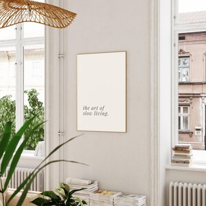 May include: A framed print with the text "the art of slow living" in a minimalist style. The print has a cream-coloured background and a thin gold frame. The image is displayed in a well-lit room with natural light.