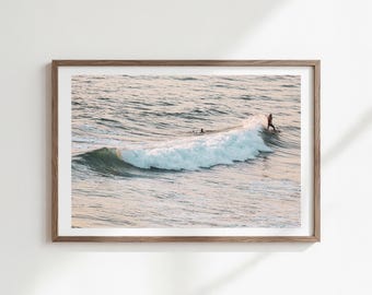 Könnte beinhalten: Gerahmter Druck mit einem Surfer, der eine Welle im Meer reitet. Die Welle bricht und erzeugt weißen Schaum. Ein weiterer Surfer ist in der Ferne zu sehen. Das Wasser ist eine Mischung aus Blau und Grau.