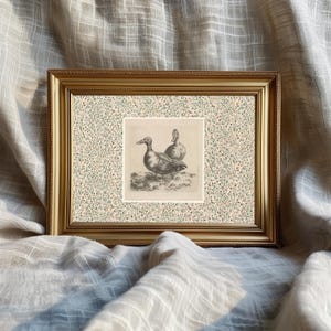 May include: A framed print featuring a detailed black and white illustration of two ducks. The artwork is set against a patterned background with a gold-colored frame. The print is displayed on a textured, neutral-toned fabric.