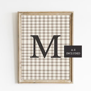 May include: Framed wall art featuring a large black letter "M" centered on a beige and white checkered background. The frame is a light brown color. A black rectangle in the lower right corner reads "A-Z INCLUDED".