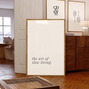 May include: A large framed print displaying the text "the art of slow living". Two other framed prints are visible, one with the word "cheers" and another with tennis rackets. The frames are a warm wood tone, creating a modern and relaxed atmosphere.
