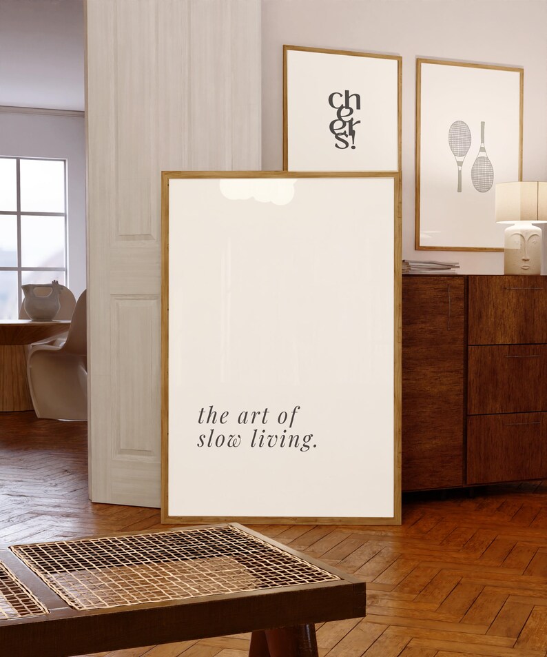 May include: A large framed print displaying the text "the art of slow living". Two other framed prints are visible, one with the word "cheers" and another with tennis rackets. The frames are a warm wood tone, creating a modern and relaxed atmosphere.