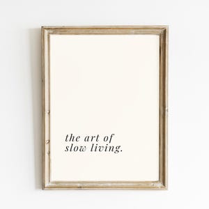 May include: A framed print featuring the phrase "the art of slow living." The print has a cream-coloured background and is set within a light brown, rustic wooden frame. The text is in a simple, elegant font.