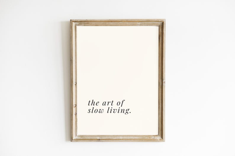 May include: A framed print featuring the phrase "the art of slow living." The print has a cream-coloured background and is set within a light brown, rustic wooden frame. The text is in a simple, elegant font.