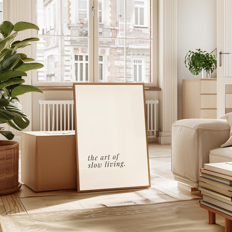 May include: A framed print with the text "the art of slow living" in a minimalist interior. The print is propped against a cardboard box, with a large window and plants in the background. The overall aesthetic is calm and inviting.