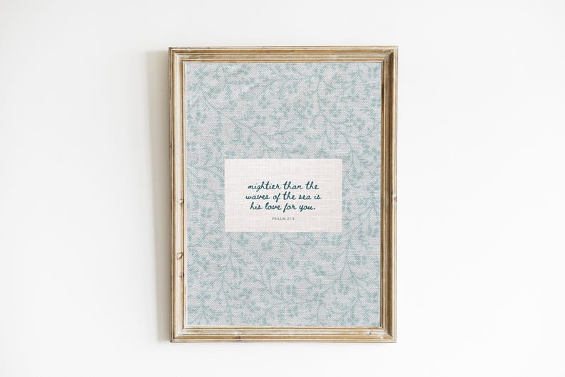Puede incluir: Arte de pared enmarcado con un fondo floral azul claro y un cuadro de texto rectangular. El cuadro de texto contiene la cita "m&aacute;s fuerte que las olas del mar es su amor por ti" en verde azulado oscuro. El marco es de color marr&oacute;n claro.